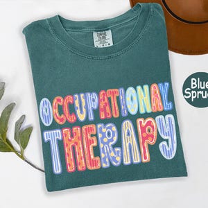 May include: A blue spruce colored t-shirt with a crew neckline and a Comfort Colors brand label. The front features the text OCCUPATIONAL THERAPY in large, multi-colored, patterned bubble letters with stripes and polka dots in pink, yellow, and blue.
