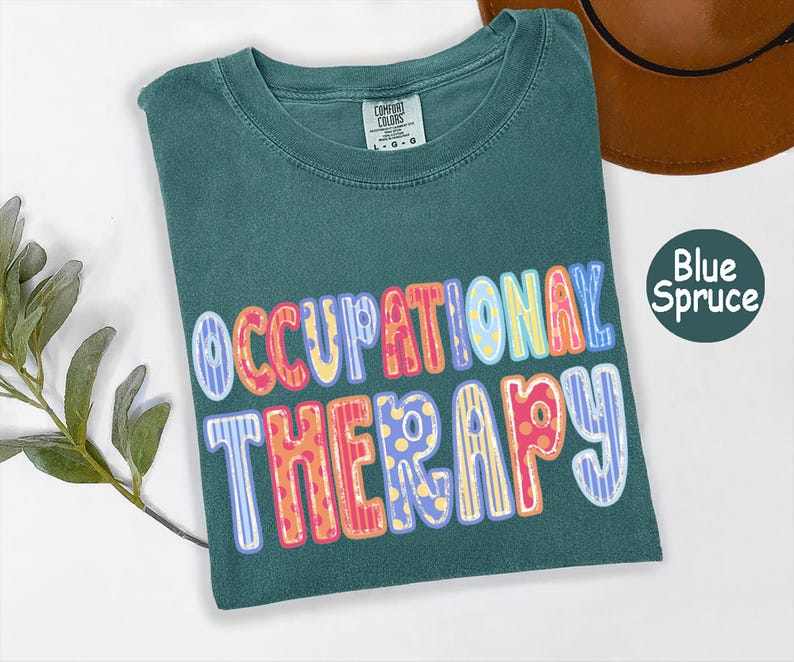 May include: A blue spruce colored t-shirt with a crew neckline and a Comfort Colors brand label. The front features the text OCCUPATIONAL THERAPY in large, multi-colored, patterned bubble letters with stripes and polka dots in pink, yellow, and blue.