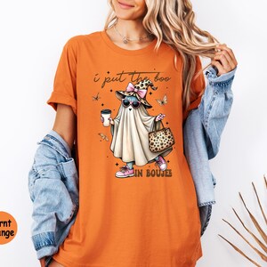 May include: A burnt orange t-shirt featuring a graphic of a ghost wearing a leopard print witch hat with a pink bow, sunglasses, and pink sneakers. The ghost holds a coffee cup and a leopard print handbag. The text reads, I put the boo in boujee.