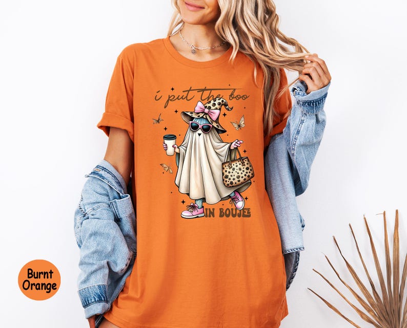 May include: A burnt orange t-shirt featuring a graphic of a ghost wearing a leopard print witch hat with a pink bow, sunglasses, and pink sneakers. The ghost holds a coffee cup and a leopard print handbag. The text reads, I put the boo in boujee.
