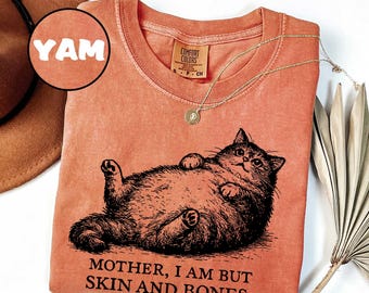Op de afbeelding: Een terracotta-kleurig t-shirt met een zwarte inkt-stijl illustratie van een dikke kat die op zijn rug ligt. Onder de kat staat de tekst: "MOTHER, I AM BUT SKIN AND BONES." Het shirt heeft een ronde hals en een Comfort Colors merklabel.