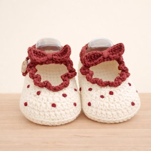 Könnte beinhalten: Ein Paar handgefertigte Babyschuhe. Die Schuhe sind cremefarben mit kleinen roten Punkten und einer roten Rüschenborte. Jeder Schuh hat eine rote Schleife und ein kleines Knopfdetail.