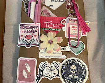 May include: A brown paper-wrapped package with stickers, pens, and a bookmark. Stickers include "Romance reader", "Trigger Warnings", and "Dark Romance Book Club". The package contains a pink pen, a white pen, and a pink tassel bookmark.