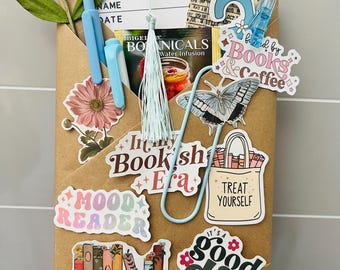 May include: A brown paper-wrapped package decorated with stickers and accessories. Stickers include "Mood Reader", "Booktrovert", and "It's a good day to read". The package also has a reading list, pens, and a paperclip. A floral arrangement and tassel add to the aesthetic.