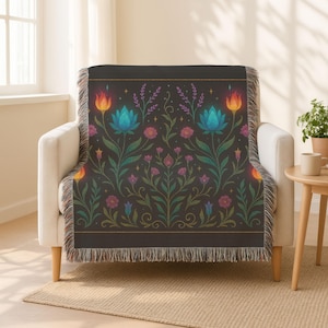 May include: A dark gray throw blanket draped over a white armchair. The blanket features a floral pattern with blue, pink, and orange flowers, green leaves, and a gold border. The blanket has a fringed edge.