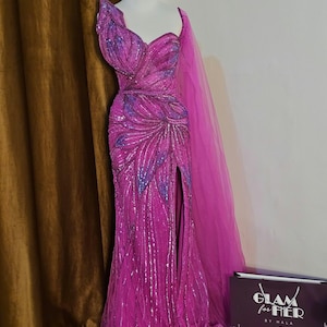 May include: A magenta-colored, floor-length gown with a high slit, embellished with sequins and intricate floral patterns. The dress features a one-shoulder design and a flowing cape. The dress is displayed on a mannequin.