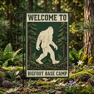 Puede incluir: Una bandera de jardín con el texto "WELCOME TO BIGFOOT BASE CAMP" y una silueta blanca de Bigfoot caminando. La bandera está situada en una escena de bosque con helechos, piñas y árboles.