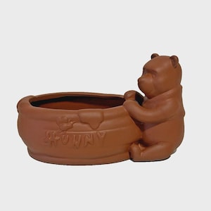 May include: A brown ceramic planter shaped like a round honey pot with a bear figure attached to the side. The pot features a relief design of a bee and the word HUNNY embossed on the front, with a smooth, matte finish and an open top.