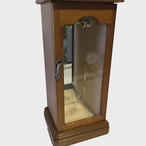 May include: A wooden jewelry cabinet with a rectangular shape and a glass door. The door features etched floral designs. The cabinet has a small handle and a decorative top. The wood has a warm, brown tone.