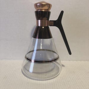May include: A vintage glass beverage server with a copper-colored lid and accents. The server has a black handle and a clear glass body with a gold-colored band. The design is a classic mid-century style.