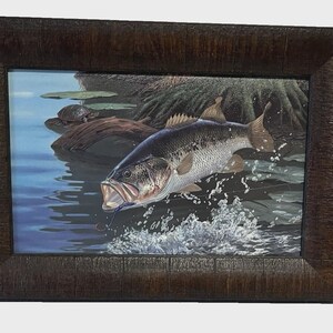 May include: A framed rectangular print featuring a largemouth bass jumping out of blue water with a fishing lure in its mouth. A small turtle rests on a log near the water's edge, and the scene is enclosed in a dark brown textured wooden frame.