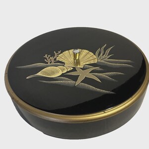 May include: A black, round trinket box with a gold-colored rim and lid. The lid features a gold seashell, starfish, and seaweed design. A small, silver-colored knob is in the center of the lid.