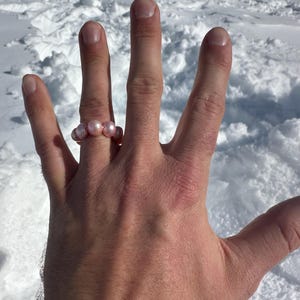 Puede incluir: Un anillo de perlas rosas en una mano. Las perlas están agrupadas para formar el anillo. El fondo es un paisaje nevado. El anillo es una joya sencilla.