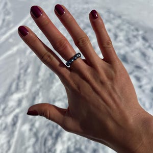 May include: A hand with burgundy nail polish displays a silver ring with five dark gray beads. The ring is worn on the ring finger. The background is a snowy, white surface.