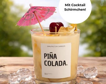 Könnte beinhalten: Ein Piña Colada-Duftkerze im Glas, hellgelb. Dekoriert mit einer Kirsche, Ananas und einem rosa Cocktail-Schirmchen. Das Etikett zeigt "PIÑA COLADA."