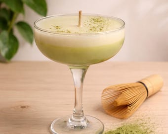 Könnte beinhalten: Eine Matcha-Kerze in einem Coupé-Glas mit einem Holzdocht. Die Kerze hat ein zweifarbiges Design mit hellgrüner Basis und cremefarbenem Oberteil, bestreut mit Matcha-Pulver. Ein Bambusbesen und Matcha-Pulver sind ebenfalls zu sehen.