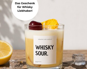 Könnte beinhalten: Eine Kerze in einem klaren Glasbehälter mit einem weißen Etikett mit der Aufschrift "JANUFACTUR CANDLES" und "WHISKY SOUR.". Die Kerze hat eine gelbe Wachsbasis, eine weiße Schicht oben, eine dunkelrote Kirsche und eine gelbe Garnitur. Der Text darüber lautet "Das Geschenk für Whisky Liebhaber!"