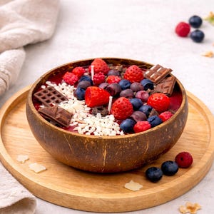May include: A decorative candle in a coconut bowl, resembling a fruit and chocolate dessert. The candle features red, blue, and white wax elements, including strawberries, blueberries, and chocolate pieces. It sits on a wooden tray with a beige cloth.