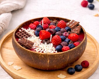 Könnte beinhalten: Dekorative Kerze in einer Kokosnussschale, die einem Obst- und Schokoladendessert ähnelt. Die Kerze hat rote Erdbeeren, blaue Blaubeeren und Schokoladenstücke. Sie steht auf einem Holztablett, im Hintergrund ist ein beiges Tuch zu sehen.