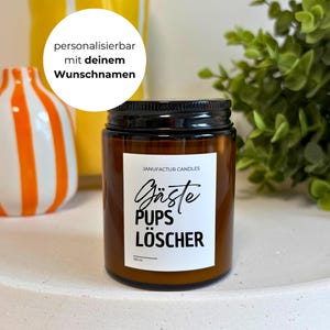 May include: A brown glass jar candle with a black lid and a white label. The label reads "Gäste Pups Löscher" in black script. The candle is on a white tray, with decorative vases in the background.