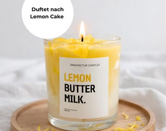 Könnte beinhalten: Eine brennende Kerze in einem transparenten Glas mit gelbem Wachs, beschriftet mit "Lemon Butter Milk". Die Kerze steht auf einem runden Holztablett, mit verstreuten gelben Wachsspänen. Der Text "Duftet nach Lemon Cake" befindet sich in einem weißen Kreis.