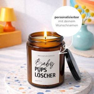 May include: A lit brown glass candle jar with a black lid, sitting on a decorative coaster. The candle has a white label with the text "Baby Pups Löscher". The candle is 150 ml.