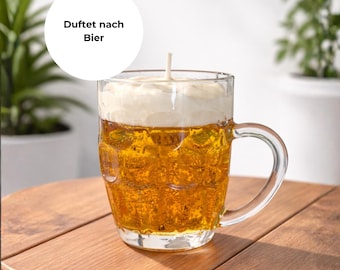 Könnte beinhalten: Eine bierförmige Kerze in einem durchsichtigen Glasbecher mit Henkel. Die Kerze hat eine goldgelbe Wachsbasis, die von einer weißen, schaumigen Schicht bedeckt ist. Der Text "Duftet nach Bier" befindet sich in einem weißen Kreis über dem Becher.
