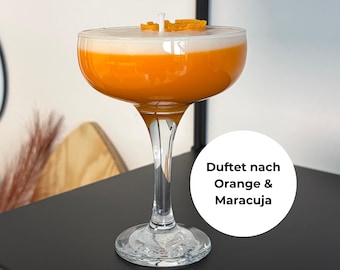 Könnte beinhalten: Eine cocktailförmige Kerze mit einem orange-weißen Wachsdesign, gekrönt von orangefarbenen Wachsstücken. Die Kerze befindet sich in einem klaren Glas mit langem Stiel und einer breiten, flachen Schale. Die Kerze steht auf einer schwarzen Oberfläche.