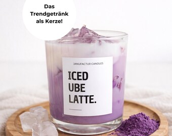 Könnte beinhalten: Eine Kerze in einem Glas, die einem Iced Ube Latte ähnelt, steht auf einem Holztablett mit Eiswürfeln und lila Pulver. Die Kerze hat ein weißes Etikett mit der Aufschrift "ICED UBE LATTE". Der Text "Das Trendgetränk als Kerze!" befindet sich in einem weißen Kreis.