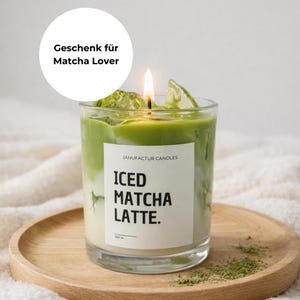 Könnte beinhalten: Eine brennende Kerze in einem Glas, das einem eisgekühlten Matcha Latte ähnelt, steht auf einem Holztablett. Die Kerze hat ein weißes Etikett mit dem Text "ICED MATCHA LATTE." und "JANUFACTUR CANDLES." Grünes Matcha-Pulver und Eiswürfel umgeben die Kerze. Der Text "Geschenk für Matcha Lover" befindet sich in einem weißen Kreis.