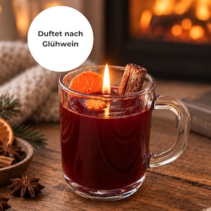 Könnte beinhalten: Ein durchsichtiger Glasbecher gefüllt mit dunkelrotem Wachs, einer brennenden Kerzendocht, einer getrockneten Orangenscheibe und einer Zimtstange. Der Text Duftet nach Glühwein steht in einem weißen Kreis über dem Becher auf einer Holzoberfläche.