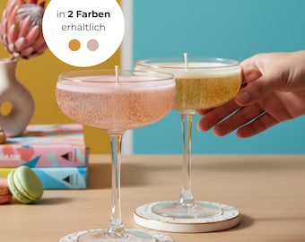 Könnte beinhalten: Zwei Champagner-Coupe-Kerzen, eine rosa und eine gelbe, stehen auf dekorativen Untersetzern. Die Kerzen haben ein sprudelndes Aussehen und einen Docht. Die Untersetzer sind weiß mit goldenem Rand und floralem Design.