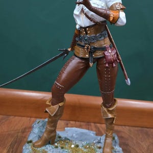 Puede incluir: Figura detallada de un personaje femenino de la serie The Witcher. Viste una parte superior blanca, pantalones de cuero marr&oacute;n y botas, con una espada a su lado. La figura se encuentra sobre una base rocosa con un emblema de lobo.