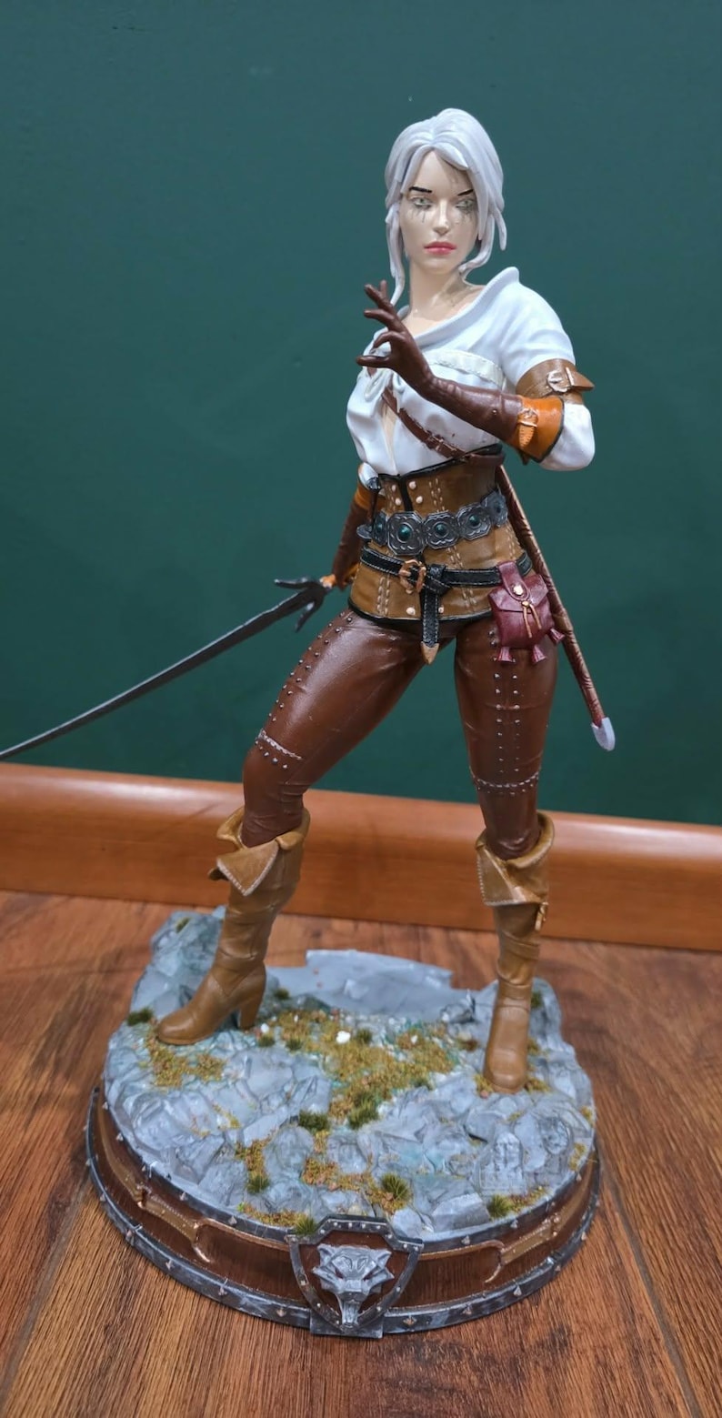 Puede incluir: Figura detallada de un personaje femenino de la serie The Witcher. Viste una parte superior blanca, pantalones de cuero marr&oacute;n y botas, con una espada a su lado. La figura se encuentra sobre una base rocosa con un emblema de lobo.