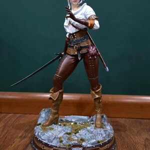 Puede incluir: Figura detallada de un personaje de la serie The Witcher. La figura viste pantalones y botas de cuero marr&oacute;n, una blusa blanca y un chaleco de cuero marr&oacute;n. Sostiene una espada y est&aacute; de pie sobre una base rocosa con un emblema de lobo.