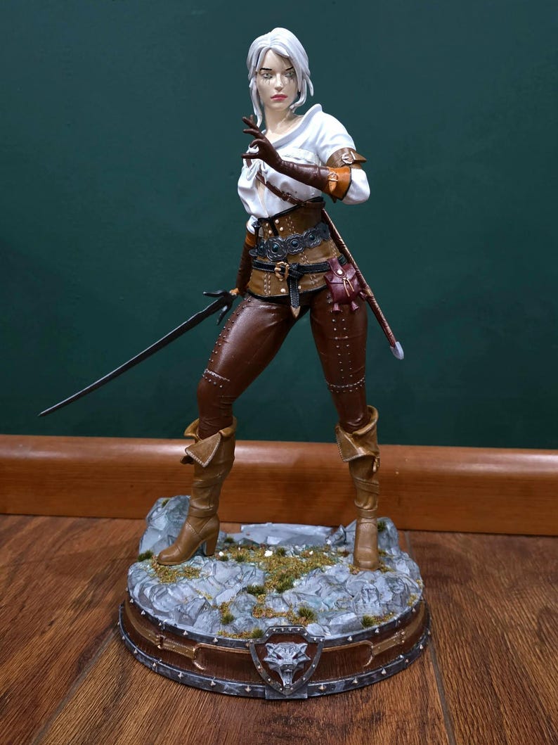 Puede incluir: Figura detallada de un personaje de la serie The Witcher. La figura viste pantalones y botas de cuero marr&oacute;n, una blusa blanca y un chaleco de cuero marr&oacute;n. Sostiene una espada y est&aacute; de pie sobre una base rocosa con un emblema de lobo.