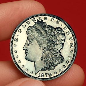 May include: A silver coin with a detailed profile of a woman, the inscriptions "E PLURIBUS UNUM" and the date "1879". The coin is held between fingers against a red backdrop. The coin's design is intricate and well-preserved.