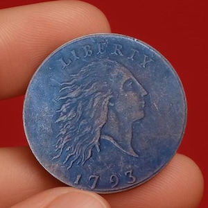 May include: A blue coin with the word "LIBERTY" and the date "1793". It features a profile of a woman with flowing hair. The coin is held between fingers against a red background. The coin's surface has a slightly aged appearance.