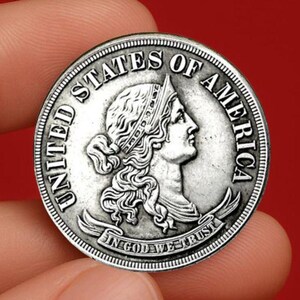 May include: A silver coin featuring a profile of a woman with a crown, surrounded by the words "UNITED STATES OF AMERICA" and "IN GOD WE TRUST". The coin is held between fingers against a red background.
