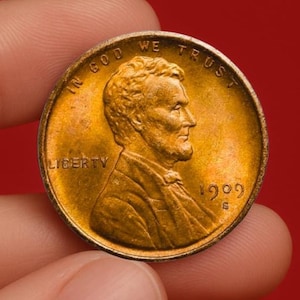 Puede incluir: Primer plano de un centavo Lincoln de 1909 sostenido entre los dedos. La moneda de cobre presenta un perfil de Abraham Lincoln, la palabra "LIBERTY" y la inscripción "IN GOD WE TRUST". La moneda es de color dorado cálido sobre un fondo rojo.