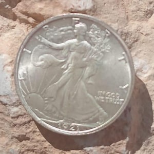 Könnte beinhalten: Eine silberne Walking Liberty Half Dollar Münze von 1921, die Lady Liberty zeigt, die auf die Sonne zugeht. Die Münze liegt auf einer strukturierten, hellbraunen Felsfläche. Die Münze trägt die Inschrift "IN GOD WE TRUST".