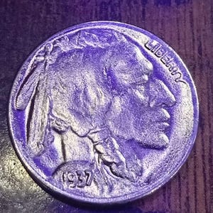 May include: A silver Buffalo nickel from 1937. The coin shows a profile of a Native American on the obverse, with the word "LIBERTY" visible. The year "1937" is also displayed. The coin is circular and has a detailed design.