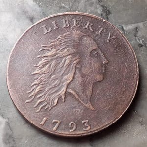 May include: A copper coin featuring a profile of a woman with flowing hair, the word "LIBERTY" above, and the date "1793" below. The coin has a worn appearance, with visible signs of age and use.