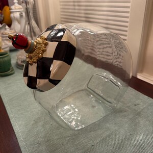 May include: A clear glass canister featuring a slanted opening and a decorative lid. The lid is painted with a black and white checkered pattern, accented by a gold-toned base and a small, round red and black plaid finial on top.