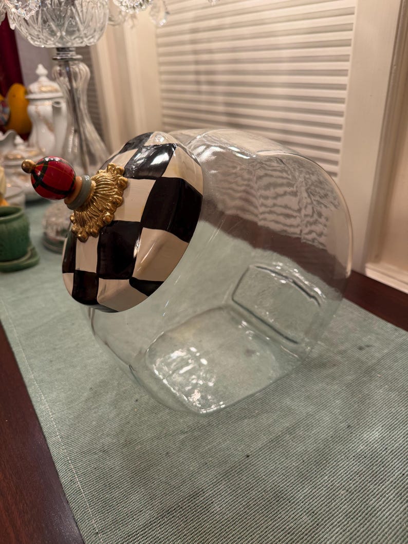 May include: A clear glass canister featuring a slanted opening and a decorative lid. The lid is painted with a black and white checkered pattern, accented by a gold-toned base and a small, round red and black plaid finial on top.