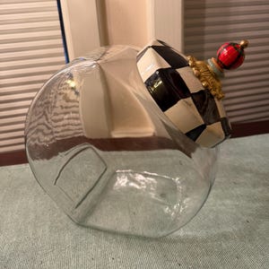 May include: A clear glass canister featuring a slanted opening and a decorative lid. The lid is adorned with a black and white checkered pattern, a gold-toned ornate base, and a small red, green, and black plaid sphere finial on top.