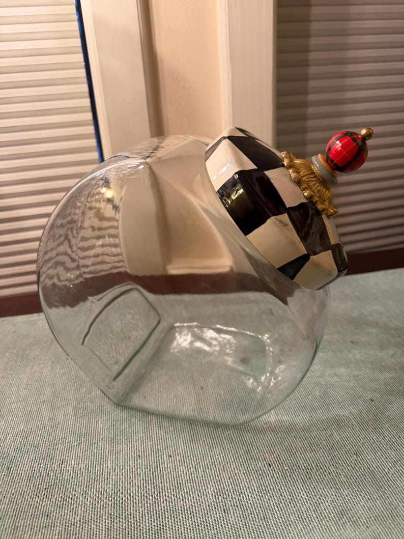 May include: A clear glass canister featuring a slanted opening and a decorative lid. The lid is adorned with a black and white checkered pattern, a gold-toned ornate base, and a small red, green, and black plaid sphere finial on top.