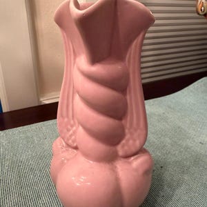 May include: A pink ceramic vase with a unique design. The vase features a twisted central column and a ruffled top. The base has a bulbous shape. The vase is a soft, pastel pink color.