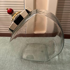 May include: A clear glass canister with a decorative lid featuring a black and white checkered pattern, a gold-toned scalloped trim, and a spherical finial painted with a red and black plaid design. The canister has a wide, rounded body shape.