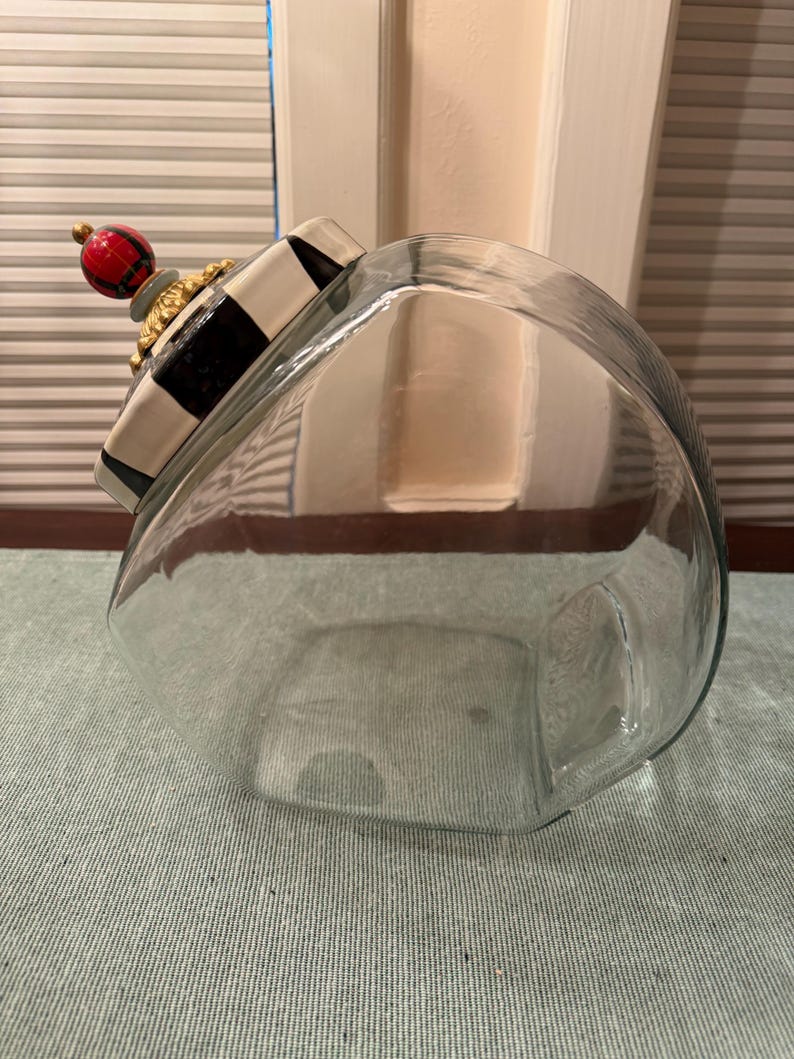 May include: A clear glass canister with a decorative lid featuring a black and white checkered pattern, a gold-toned scalloped trim, and a spherical finial painted with a red and black plaid design. The canister has a wide, rounded body shape.